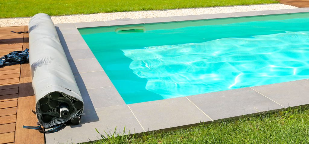 Description pour la balise alt SEO : "Piscine rectangulaire moderne avec couverture de sécurité enroulée sur un côté, installée sur une terrasse en bois et carrelage avec pelouse verte en arrière-plan."