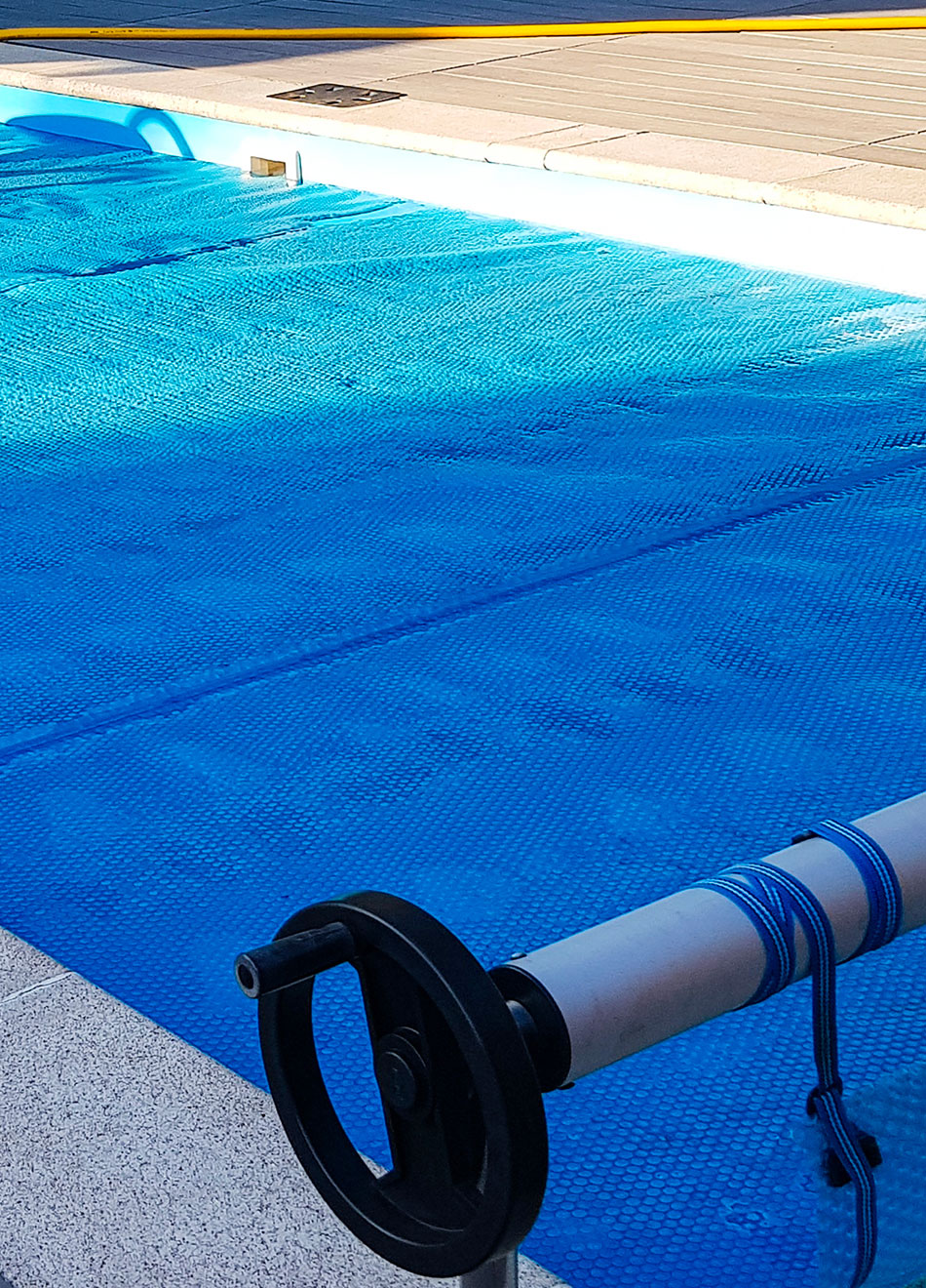 Couverture de piscine bleue partiellement enroulée sur un mécanisme de bobinage près du bord de la piscine.