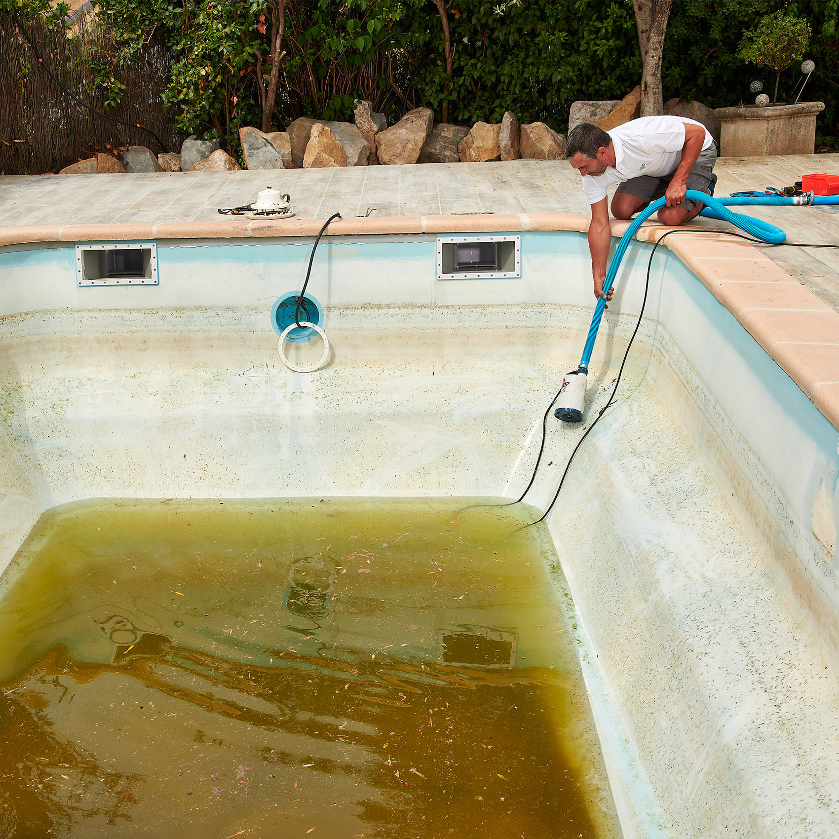 Homme nettoyant une piscine sale avec un aspirateur manuel, entouré de rochers et de végétation.