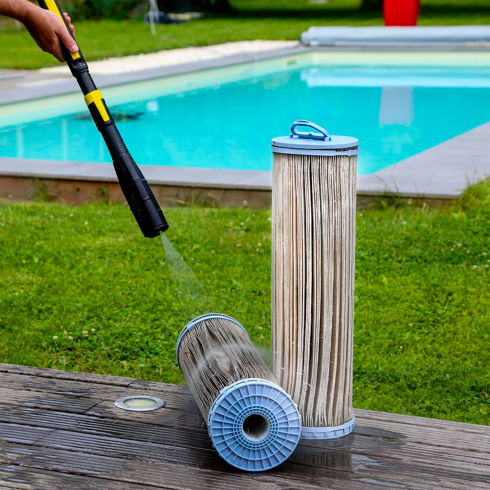 Nettoyage d'un filtre de piscine avec un nettoyeur haute pression sur une terrasse en bois, avec une piscine en arrière-plan.