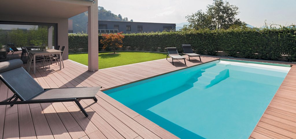 Terrasse en bois moderne avec piscine, chaises longues et salle à manger extérieure, entourée d’une haie verte.