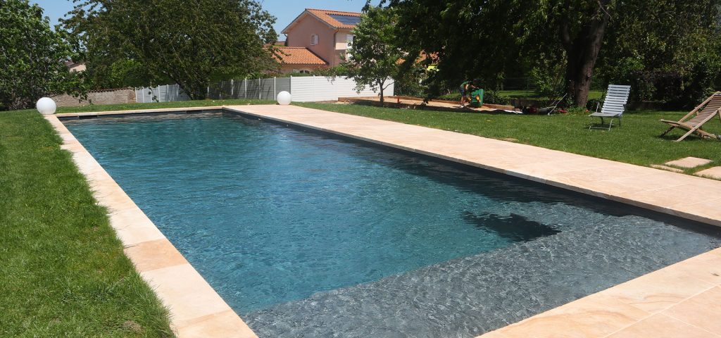 Piscine rectangulaire en plein air avec bordures en pierre claire et pelouse verdoyante, dans un jardin résidentiel spacieux.