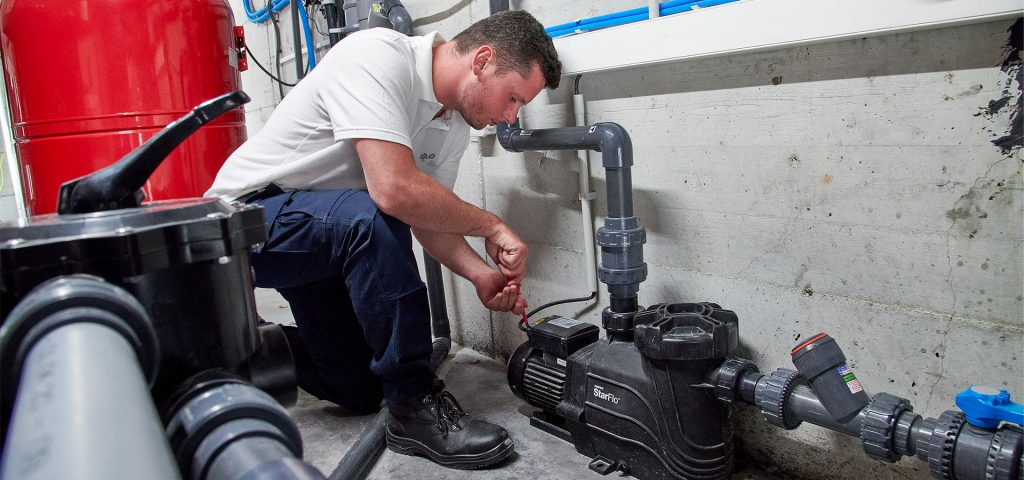 Technicien en uniforme réparant une pompe à eau dans une salle des machines avec des conduits et une grande citerne rouge.