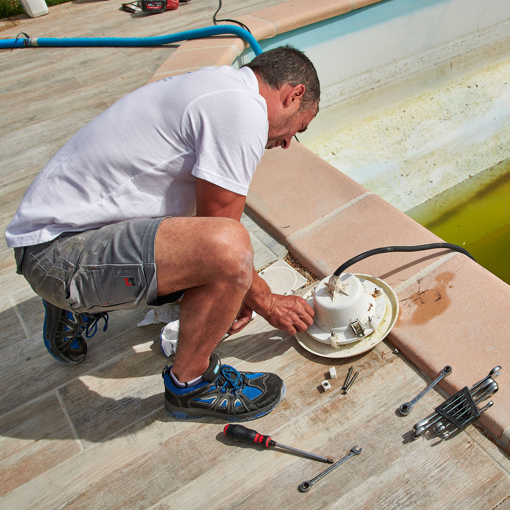 Homme réparant une pompe de piscine avec des outils à côté sur une terrasse ensoleillée.