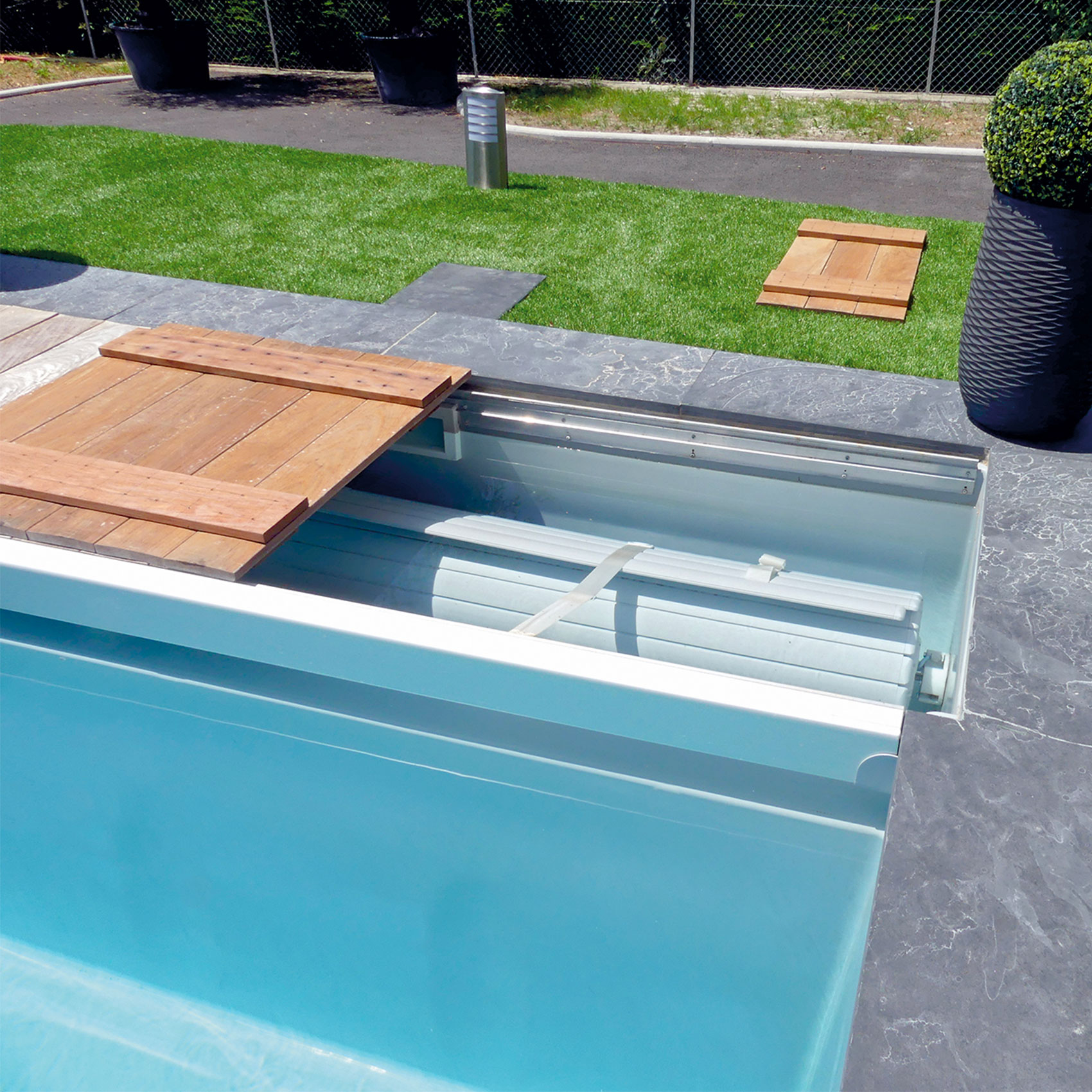 Couverture de piscine automatique intégrée ouverte sur une piscine extérieure avec des dalles en pierre et un gazon artificiel en arrière-plan.