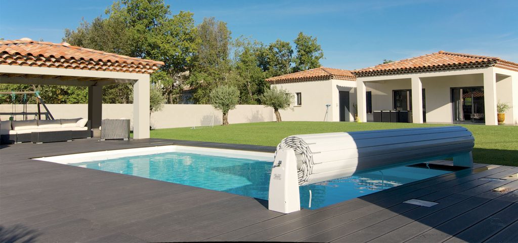 Maison moderne avec piscine, terrasse en bois, couverture de piscine automatique, et zone de relaxation sous pergola dans une cour verdoyante.