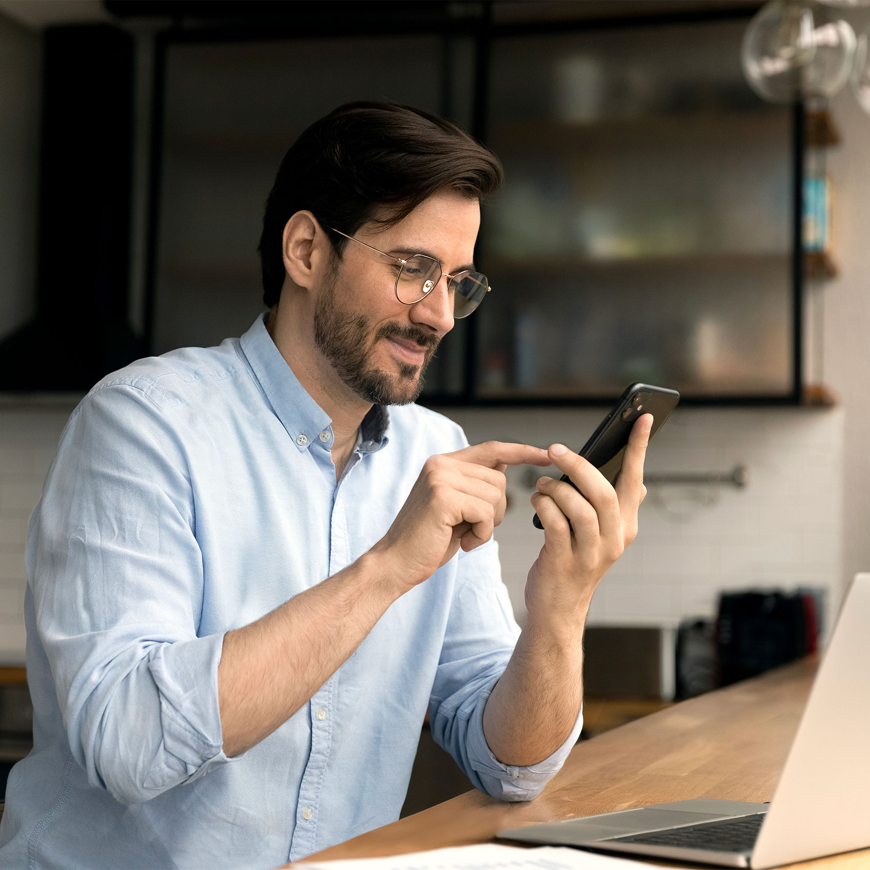 Homme souriant utilisant son smartphone dans un bureau moderne avec un ordinateur portable.