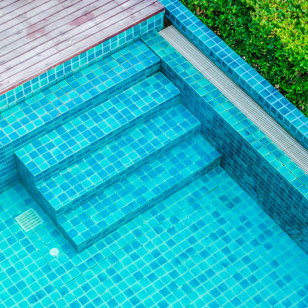 Escaliers de piscine en mosaïque bleue avec rebord en bois et haie verte en fond.