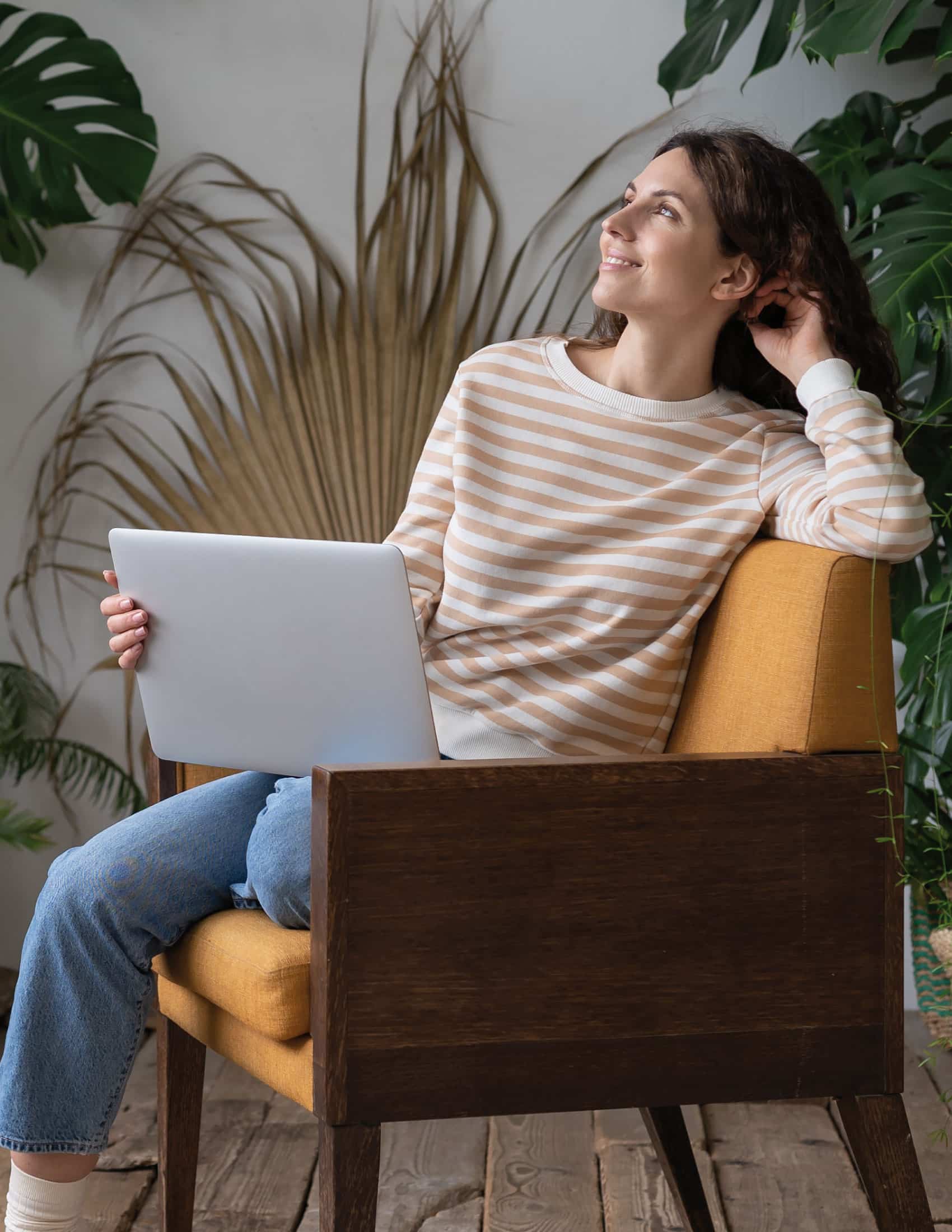 Femme souriante en jeans assise dans un fauteuil moutarde tenant un ordinateur portable, entourée de plantes d'intérieur.